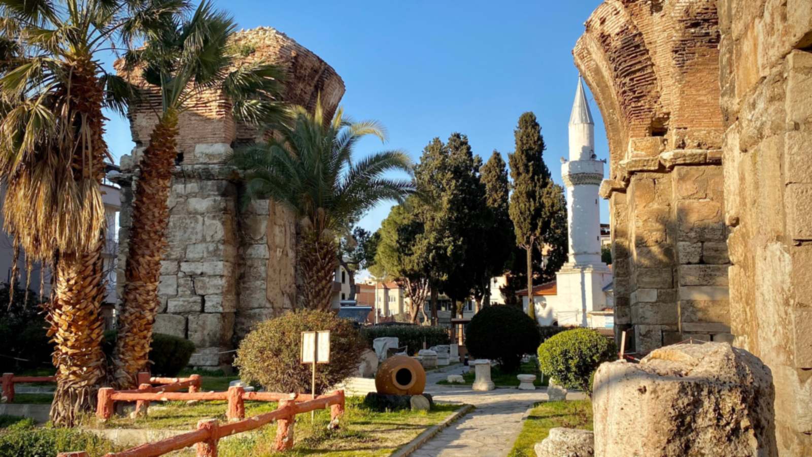 Brick-and-stone pillars of the Basilica of St. John in Alaşehir, ancient Philadelphia, with the white minaret of a neighborhood mosque beyond the garden